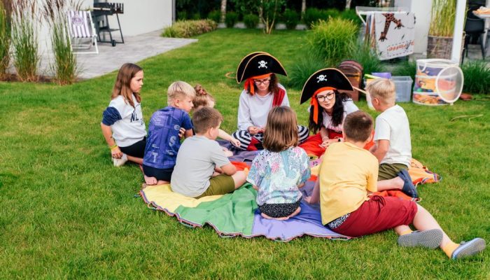 Dzieci bawią się na trawie wokół kolorowego koca, uczestnicząc w zabawie z postaciami przebranymi za piratów. Scena przedstawia radosne chwile w luksusowym domku nad morzem w Władysławowie, idealnym na wakacje z dziećmi. Domki oferują atrakcje dla rodzin, w tym plac zabaw, co sprawia, że są doskonałym miejscem na weekend z dziećmi oraz letnie wyjazdy nad morzem.