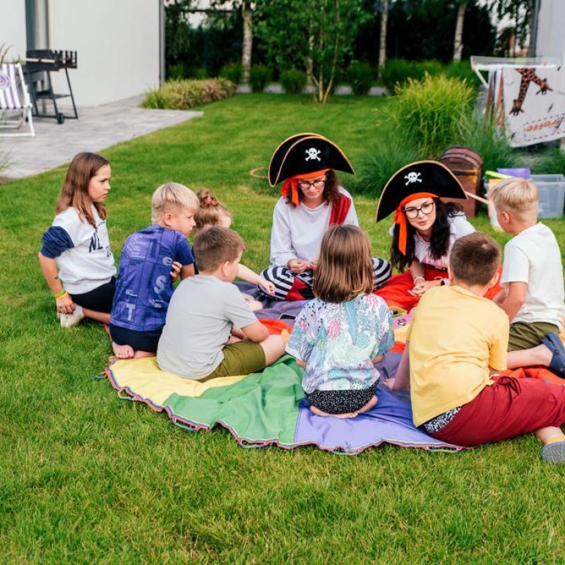 Dzieci bawią się na trawie wokół kolorowego koca, uczestnicząc w zabawie z postaciami przebranymi za piratów. Scena przedstawia radosne chwile w luksusowym domku nad morzem w Władysławowie, idealnym na wakacje z dziećmi. Domki oferują atrakcje dla rodzin, w tym plac zabaw, co sprawia, że są doskonałym miejscem na weekend z dziećmi oraz letnie wyjazdy nad morzem.